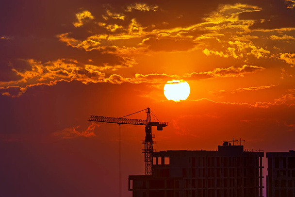 Black silhouettes of buildings under construction and crane against the sun and orange sunset, evening in the city, dramatic cloudy sky. Yerevan, Armenia. Perfect colorful background for a text - Photo, Image