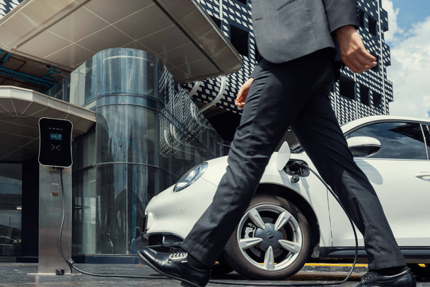 Closeup progressive businessman with electric car recharging at public charging station with background of city residential building. Eco friendly car powered by alternative clean energy. - 写真・画像