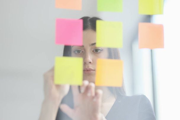 Brunette woman puts sticky notes on glass wall in modern office. Focused female manager generates new ideas and plan for business project view through glass - Photo, Image