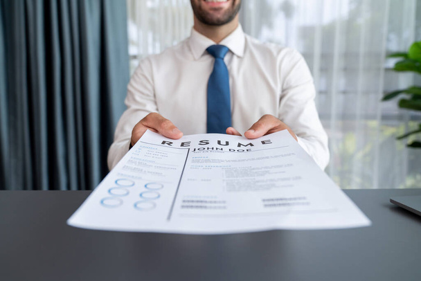 Confident man wearing suit in formal office, hand holding resume paper during job interview. Interviewer point of view with candidate handing resume paper in front of camera for consideration. Fervent - Photo, Image
