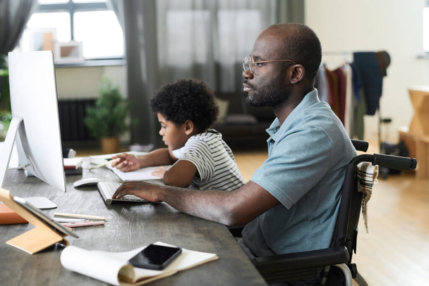 Afrikanischer behinderter Vater arbeitet online am Computer und sitzt im Rollstuhl am Schreibtisch, während sein Sohn im Hintergrund Hausaufgaben macht - Foto, Bild