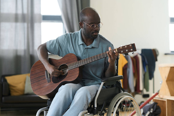 Uomo afroamericano in occhiali seduto sulla sedia a rotelle e imparare a suonare la chitarra durante il suo tempo libero a casa - Foto, immagini