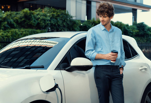 Progressive eco-friendly concept of parking EV car at public electric-powered charging station in city with blur background of businessman leaning on recharging-electric vehicle with coffee. - Photo, image