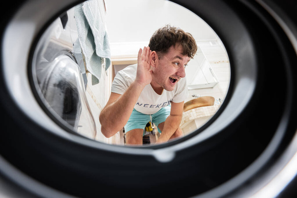 Вид чоловіка з пральної машини всередині. Чоловік займається щоденною пральнею. Слухає звук всередині
. - Фото, зображення