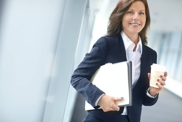 Cheerful businesswoman holding folders and drinking a coffee at work. One female businessperson carrying files and drinking tea on a break in an office. Business professional at a startup company. - Photo, Image