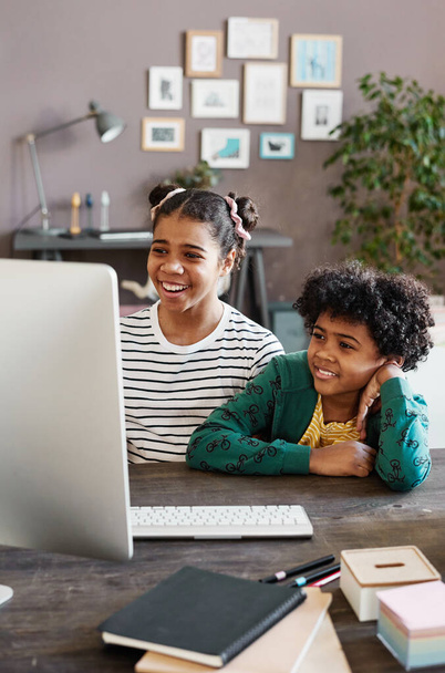 Szczęśliwy brat i siostra siedzi przy stole w pokoju przed monitorem komputera i uśmiecha się podczas rozmowy wideo - Zdjęcie, obraz