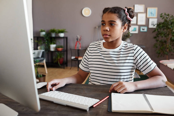 Африканська школярка сидить за столом з записником і дивиться на монітор комп "ютера. - Фото, зображення