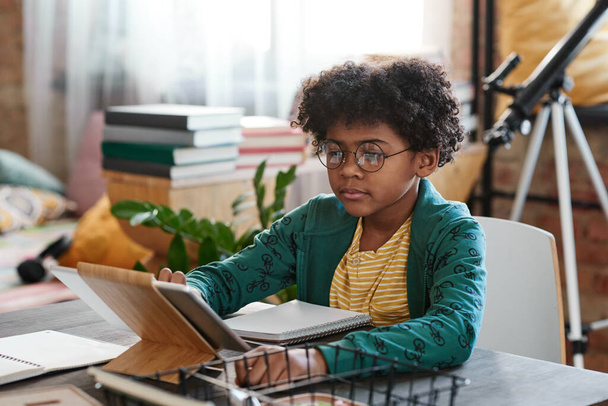 Niño africano con el pelo rizado en gafas tocando la pantalla de la tableta PC para hacer la tarea en línea en la mesa con portátil - Foto, imagen