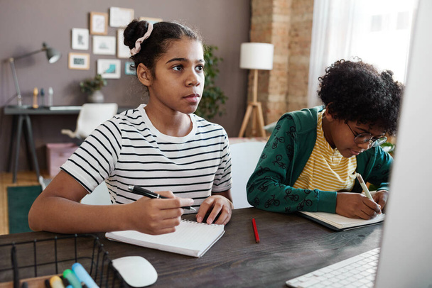 Dvě děti z africké školy sedící u stolu před monitorem počítače a zapisující si poznámky do sešitů během online lekce - Fotografie, Obrázek