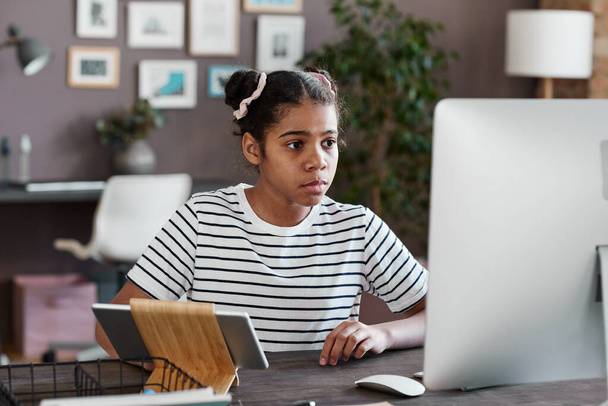 Afrikkalainen tyttö istuu pöydässään tietokoneen näytöllä ja keskittyy hänen pelata videopeli - Valokuva, kuva
