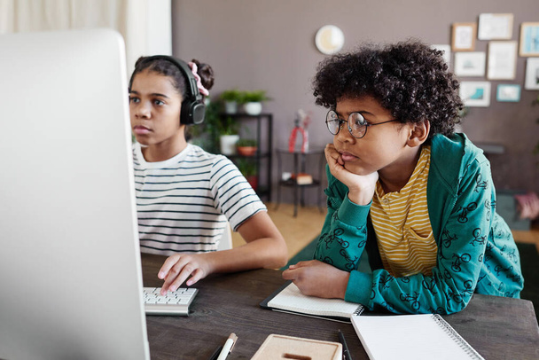 African boy in eyeglasses looking at computer monitor while girl playing video game on computer during their leisure time at home - Photo, Image