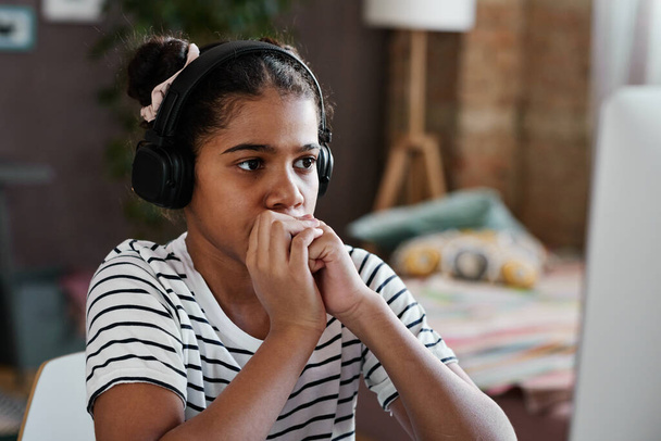 Africana chica seria en auriculares inalámbricos sentado frente a la computadora en casa y mirando el monitor - Foto, Imagen