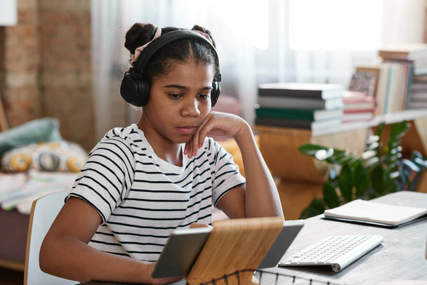 Afričanky dívka v bezdrátových sluchátkách sedí u stolu a sledovat na obrazovce digitální tablet během videohovoru - Fotografie, Obrázek
