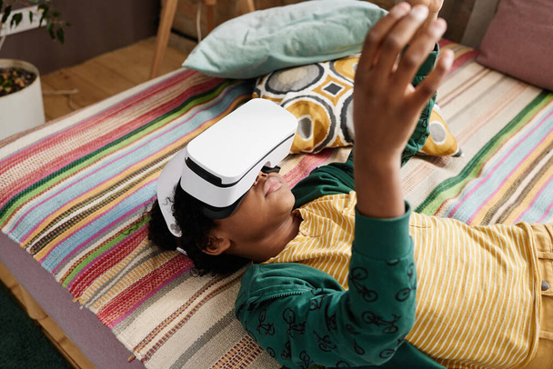Niño africano acostado en el sofá en gafas VR jugando juego de realidad virtual y gestos - Foto, Imagen