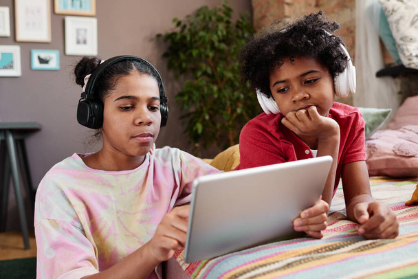 Irmão e irmã em fones de ouvido sem fio assistindo vídeo online em tablet digital juntos em casa - Foto, Imagem