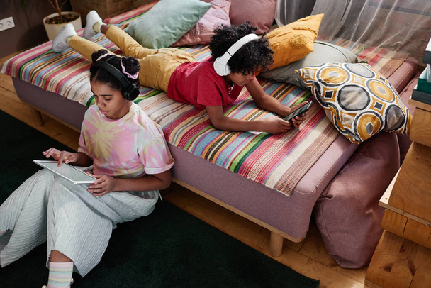 Vista ad alto angolo dei bambini africani in cuffia che trascorrono il loro tempo libero per gadget, giocando e guardando video online nella stanza - Foto, immagini