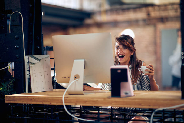 Joyful young woman looking at screen holding coffee, sitting in the office feeling excited. Business, design, freelance concept. - Photo, Image