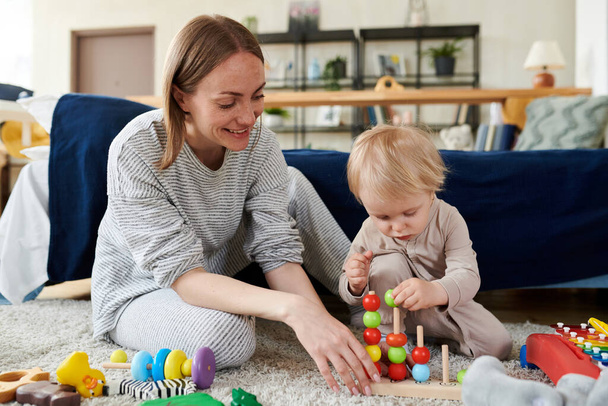 Sorrindo jovem mãe sentada no chão e assistindo por seu filhinho jogando brinquedos educativos - Foto, Imagem