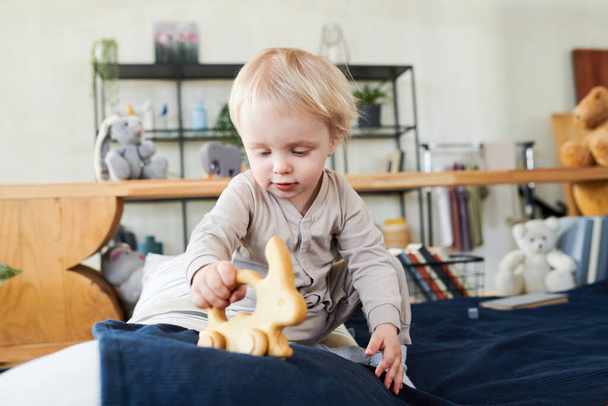 Kleiner Junge mit blonden Haaren spielt in seiner aktiven Zeit mit seinem Lieblingsspielzeug auf dem Bett - Foto, Bild