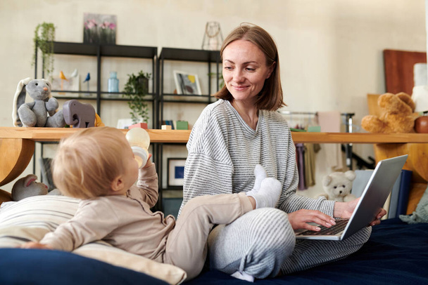 Młoda matka pracuje online na laptopie, podczas gdy jej dziecko pije wodę z butelki, siedzą na łóżku w pokoju - Zdjęcie, obraz