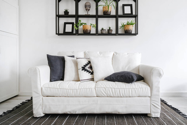 interior bohemio en sala de estar, vista frontal en sofá confort con cojines en fundas de algodón, estante de metal negro con decoración y plantas en maceta en la pared y alfombra en el suelo - Foto, Imagen