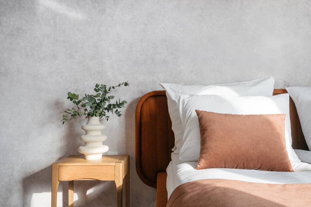 az eukaliptusz ág közelsége kerámia vázában, fa éjjeliszekrényen, kényelmes ágy mellett párnákkal, ágyneműkkel és barna bársonyborítással a háttérben a hotelszobában - Fotó, kép