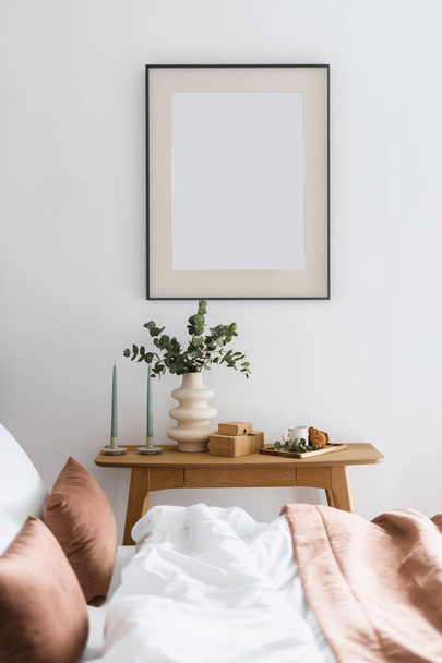 näkymä sängyn läpi tyynyt ja vuodevaatteet tyhjä taide juliste runko seinälle, kynttilät, keraaminen maljakko eukalyptus haara, kuppi ja croissant puinen pöytä moderni makuuhuone - Valokuva, kuva