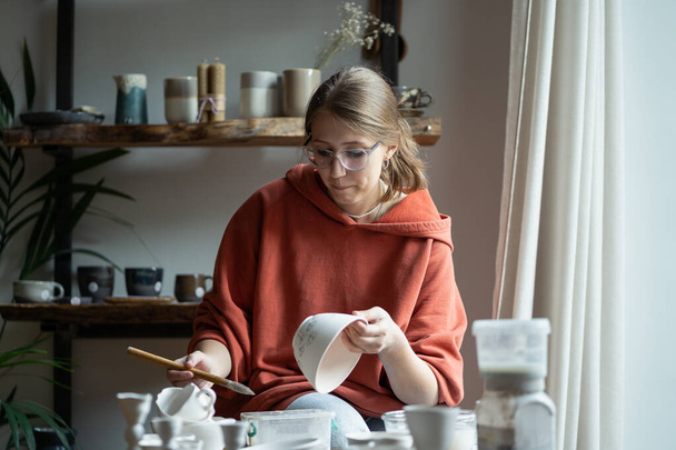 Concentrated creative girl student from art college sits at table in workshop doing coloring dishes. Thoughtful woman getting rid of stress by making handmade tableware in home handicraft studio  - Fotografie, Obrázek