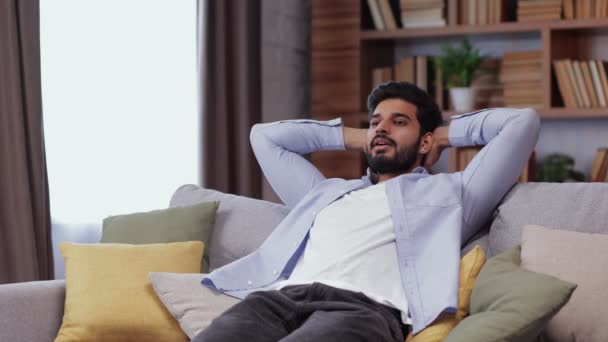 Ηρεμία χαλαρή ινδική νεαρός αναπαύεται στον καναπέ στο σπίτι, τα χέρια πίσω από το κεφάλι απολαμβάνουν το άγχος ελεύθερη ειρηνική ημέρα. Ο νεαρός Ινδός νιώθει καλά χαμογελώντας.  - Πλάνα, βίντεο