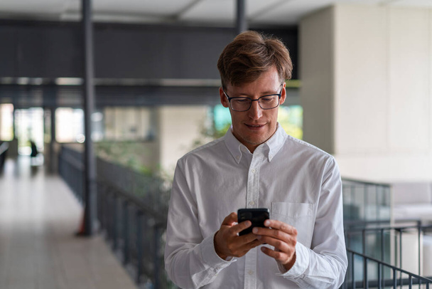 Smiling young European businessman in glasses using smartphone in blurry office hall. Concept of communication and social media - Foto, immagini