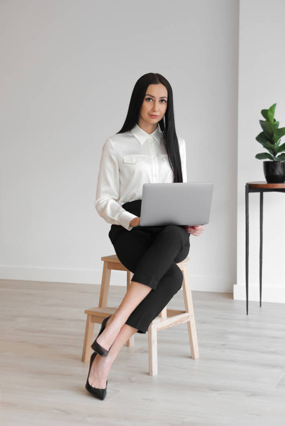 Full length shot of a serious elegant pretty businesswoman executive with long hair, sitting on a stool, using a laptop, looking at camera, at workplace. Vertical , - Fotoğraf, Görsel