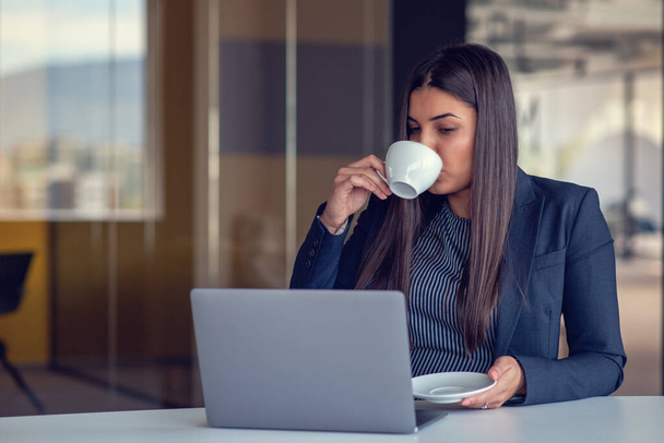 Young woman working on laptop and drinking coffee in office. - Фото, зображення
