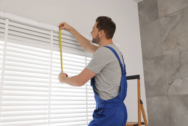 Worker in uniform using measuring tape while installing horizontal window blinds indoors - Foto, imagen