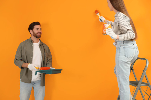Woman painting orange wall and man holding container with roller. Interior design - 写真・画像