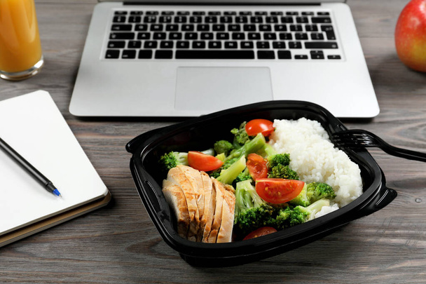 Container with tasty food, notebook and laptop on wooden table. Business lunch - Φωτογραφία, εικόνα