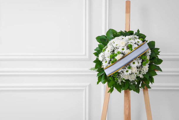 Funeral wreath of flowers with ribbon on wooden stand near white wall indoors. Space for text - Photo, Image