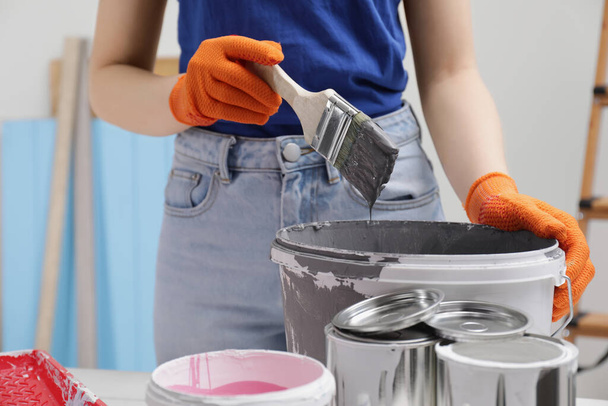 Woman dipping brush into bucket of grey paint at table indoors, closeup - Foto, Bild