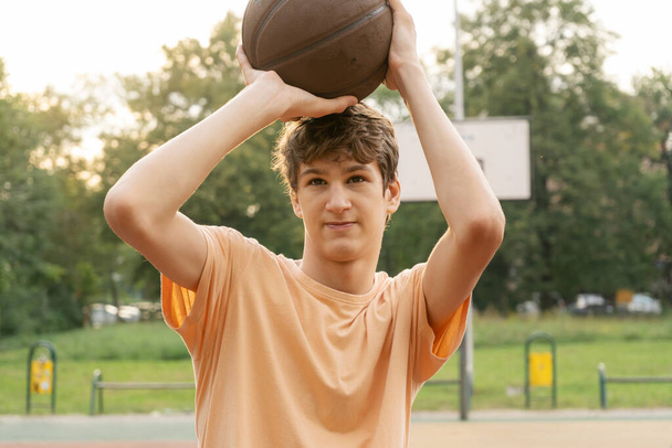 Cute young teenager in t shirt with a ball plays basketball on court. Teenager running in the stadium. Sports, hobby, active lifestyle for boys - Zdjęcie, obraz