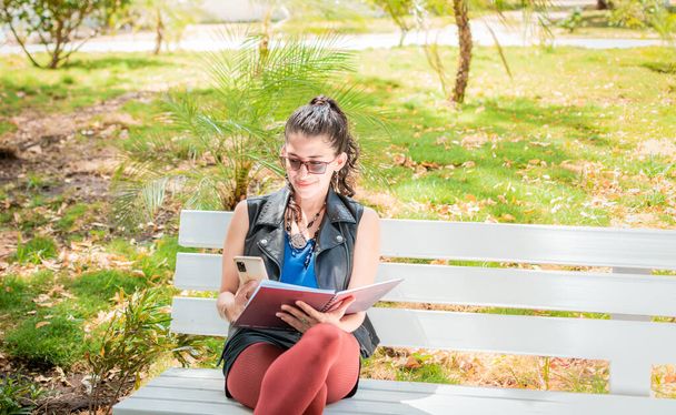 A girl sitting taking notes with a notebook and cell phone, Woman sitting taking notes with her cell phone, Latin girl using her cell phone in a park - Photo, image