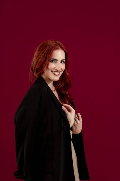 Close-up de uma mulher de cabelos vermelhos confiante olhando para a câmera enquanto posando contra um fundo isolado vermelho. Quadro vertical. - Foto, Imagem