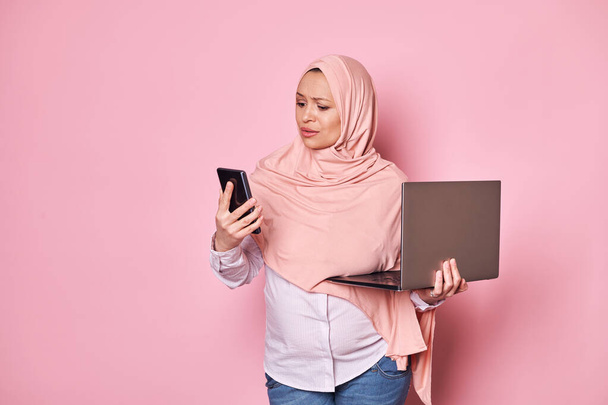 Ratlos schockierte arabische Muslimin, die im rosafarbenen Hijab arbeitet, mit verblüfftem Blick auf ihr Smartphone blickt, Missverständnisse äußert, mit Laptop vor rosafarbenem Hintergrund posiert - Foto, Bild