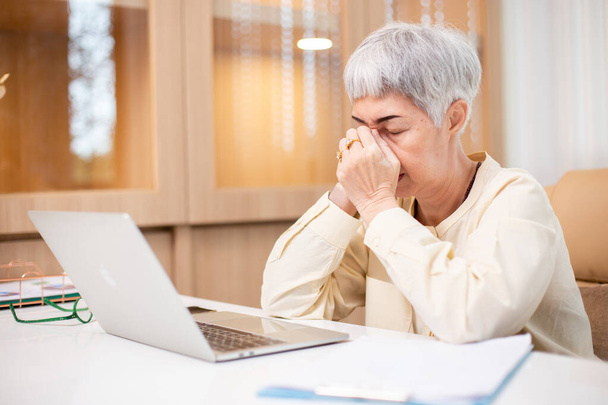 Working in retirement concept, Asian senior business woman stress from hard work, Female manager headache sitting at table with laptop. Worried middle aged female health problems. - Photo, Image