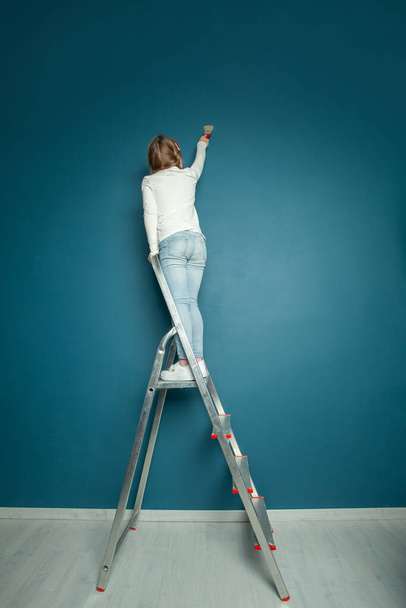Girl painting blue wall, doing renovation with brush and standing on the ladder - Foto, Imagem