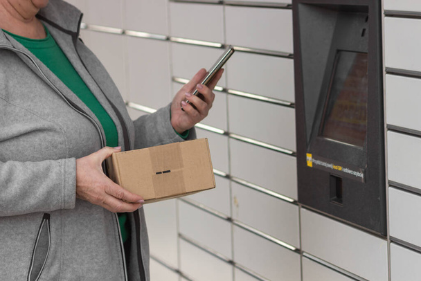 Pniewy, Polonya - 22 Mart 2023. Akıllı telefonu olan yaşlı bir kadın posta servisindeki alışveriş dolabını açmak için şifre giriyor ya da bir kutu gönderiyor. E-ticaret nakliye ve teslimat konsepti. - Fotoğraf, Görsel