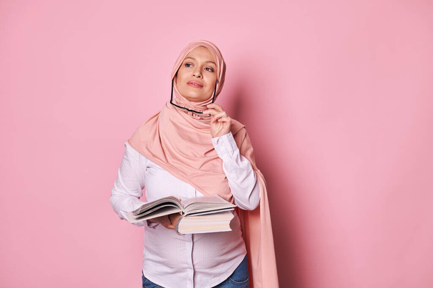 Pensive Arab Muslim pregnant woman in eyeglasses and pink hijab, holding studying book, dreamily looking away, isolated on pink background. Pretty female reasoning on her plans while maternity leave - Фото, зображення