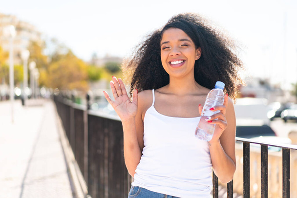 Молодая афроамериканка с бутылкой воды на открытом воздухе салютует со счастливым выражением лица - Фото, изображение