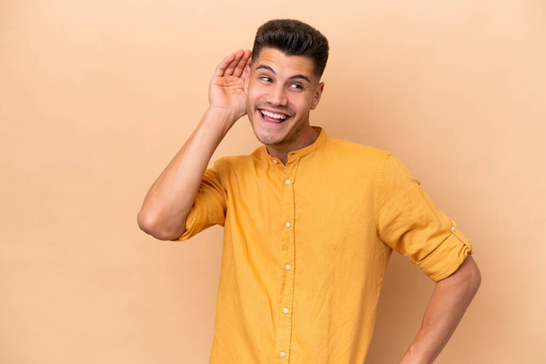 Young caucasian man isolated on beige background listening to something by putting hand on the ear - Photo, Image