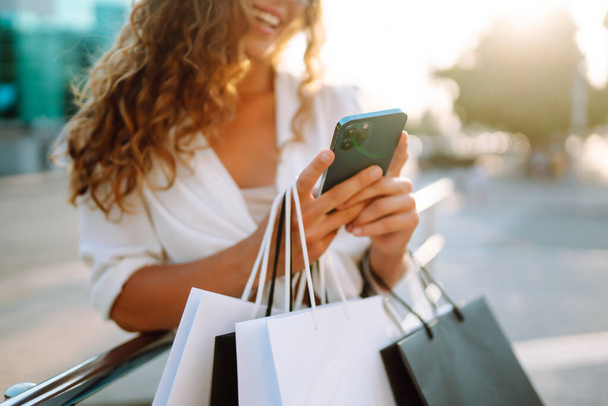 Stylish young woman shopping with a phone on a sunny street. Beautiful woman is using mobile phone and holding shopping bags. Concept of shopping, selling, consumerism. Active lifestyle. - Foto, imagen
