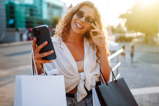 Stylish young woman shopping with a phone on a sunny street. Beautiful woman is using mobile phone and holding shopping bags. Concept of shopping, selling, consumerism. Active lifestyle. - Zdjęcie, obraz