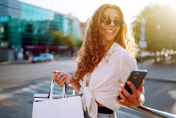 Stylish young woman shopping with a phone on a sunny street. Beautiful woman is using mobile phone and holding shopping bags. Concept of shopping, selling, consumerism. Active lifestyle. - Foto, Imagem